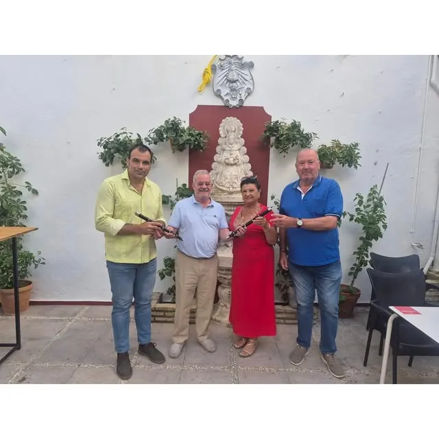 Cuatro personas posan en un patio con plantas y una imagen religiosa sosteniendo txistus donados a la Escuela de Tamborileros de la Hermandad