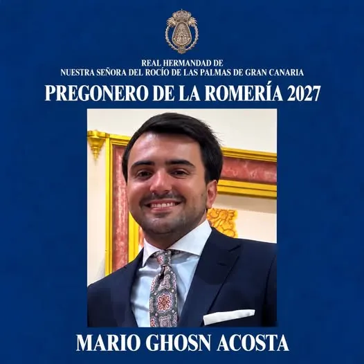Hombre joven sonriente con traje oscuro, camisa blanca y corbata estampada posando frente a fondo interior con marco dorado