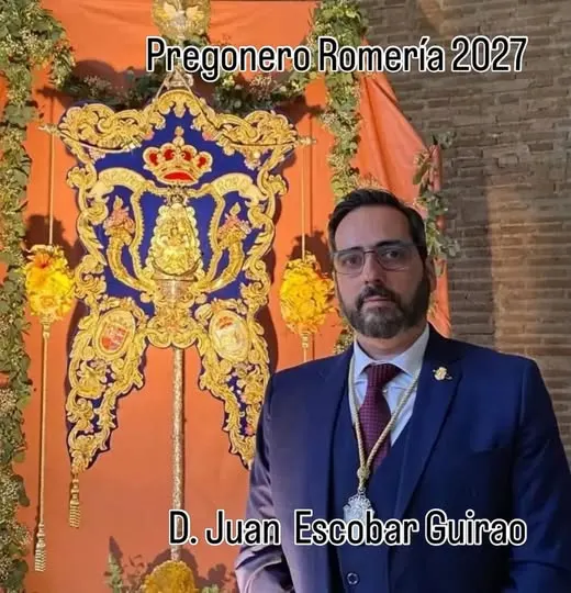 Hombre con traje azul y corbata roja posa junto a un estandarte rociero azul y dorado con corona y flores amarillas en fondo naranja