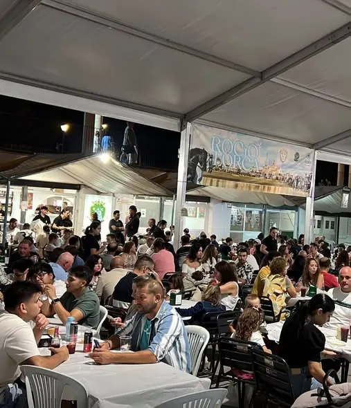 Personas de todas las edades sentadas en mesas bajo una carpa disfrutando de comida y bebida en la caseta de la Hermandad del Rocío de Lorca durante la feria nocturna