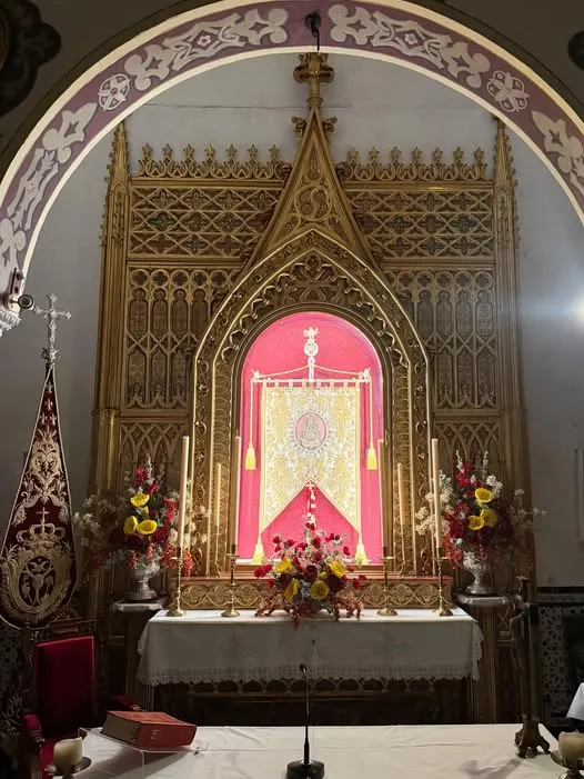 Retablo gótico dorado iluminado con luz cálida que resalta un estandarte central rojo y dorado, flanqueado por arreglos florales y velas en la ermita de Roncesvalles