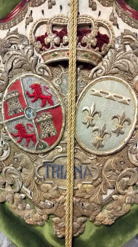 Bordado detallado en tonos dorados y rojos con escudos, corona real y la palabra Triana sobre fondo verde en estandarte religioso