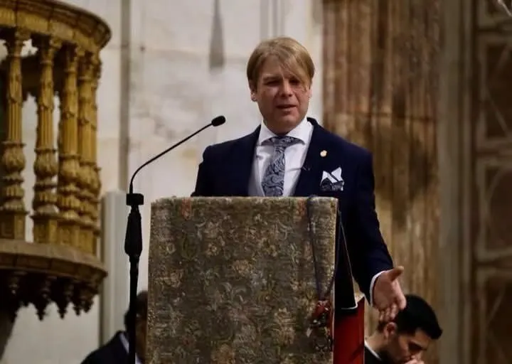Hombre vestido con traje oscuro y corbata clara hablando en un atril decorado dentro de un edificio histórico con columnas talladas y paredes de piedra clara