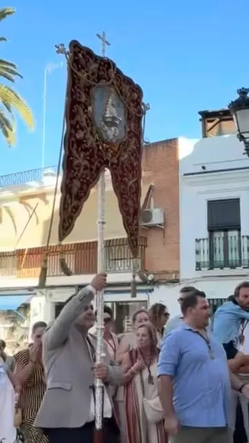 Hombre sostiene estandarte religioso con fondo de edificios y grupo de personas en una calle durante peregrinación diurna