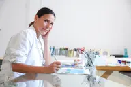 Mujer con bata blanca pintando en una mesa de cristal en un estudio con pinceles y materiales de pintura al fondo