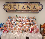 Muñecos vestidos con ropajes tradicionales y etiquetas con nombres, dispuestos en filas bajo un azulejo con la palabra TRIANA en letras grandes y azules