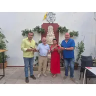 Cuatro personas posan en un patio con plantas y una imagen religiosa sosteniendo txistus donados a la Escuela de Tamborileros de la Hermandad