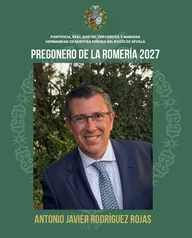 Hombre con gafas, traje azul y corbata clara sonríe frente a un fondo de árboles en un cartel verde que anuncia su nombramiento como pregonero de la Romería 2027