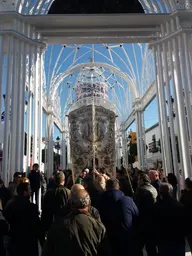 Grupo de personas reunidas bajo una estructura blanca metálica con un estandarte bordado en el centro durante una procesión religiosa en día soleado