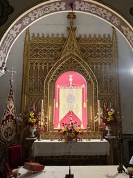 Retablo gótico dorado iluminado con luz cálida que resalta un estandarte central rojo y dorado, flanqueado por arreglos florales y velas en la ermita de Roncesvalles