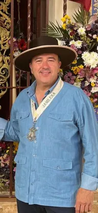 Hombre sonriente con sombrero tradicional y camisa azul lleva medalla colgada con cinta que dice Jamilena, posando frente a altar floral con flores variadas y fondo decorado