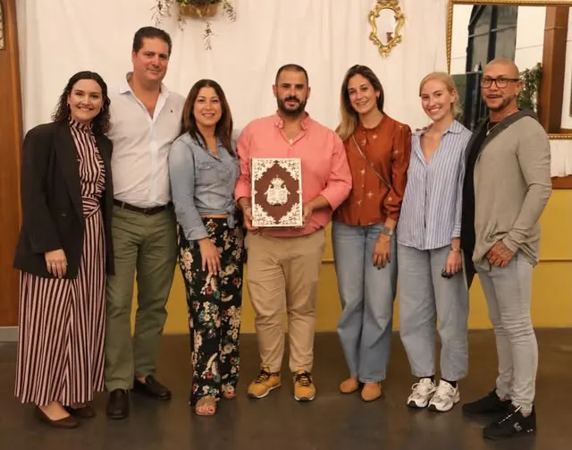 Siete personas posan en fila en un interior, el hombre del centro sostiene un libro con portada decorada mientras todos sonríen y visten ropa casual