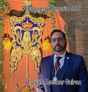 Hombre con traje azul y corbata roja posa junto a un estandarte rociero azul y dorado con corona y flores amarillas en fondo naranja
