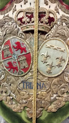 Bordado detallado en tonos dorados y rojos con escudos, corona real y la palabra Triana sobre fondo verde en estandarte religioso