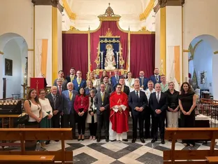 Grupo de personas posando en el interior de una iglesia frente a un altar con un retablo dorado y cortinas rojas, en un acto solemne cofrade