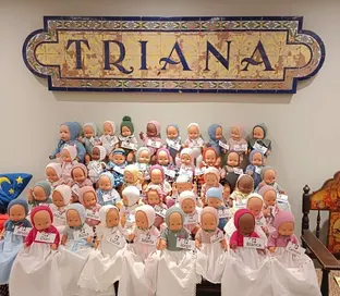 Muñecos vestidos con ropajes tradicionales y etiquetas con nombres, dispuestos en filas bajo un azulejo con la palabra TRIANA en letras grandes y azules