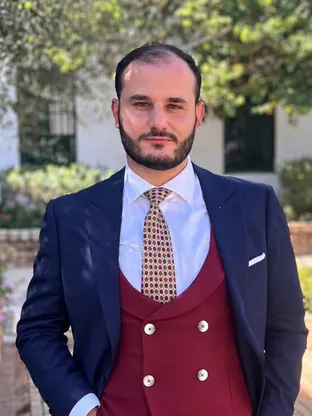 Hombre con traje azul marino, chaleco burdeos y corbata de motivos geométricos posa al aire libre con fondo de árboles y muro blanco
