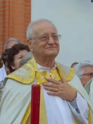 Hombre mayor con vestimenta litúrgica blanca y dorada, gafas y expresión serena, rodeado de personas en un acto religioso al aire libre