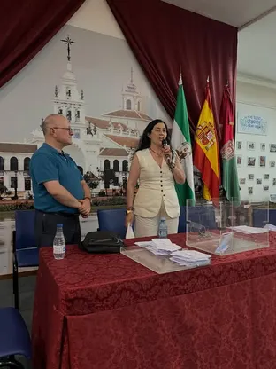 Dos personas de pie junto a una mesa con urnas transparentes y papeletas en un salón con banderas y un gran mural de un edificio histórico al fondo