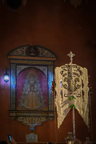 Simpecado iluminado en primer plano dentro de la Parroquia de San Jacinto con un cuadro de la Virgen del Rocío al fondo y fieles presentes en la ceremonia