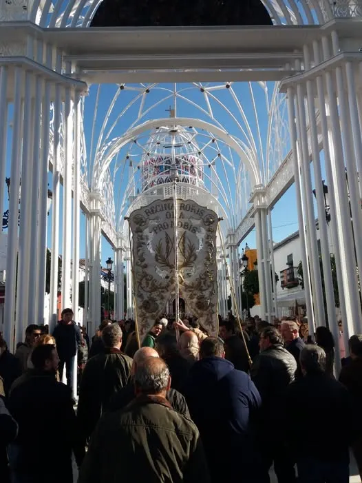 Grupo de personas reunidas bajo una estructura blanca metálica con un estandarte bordado en el centro durante una procesión religiosa en día soleado