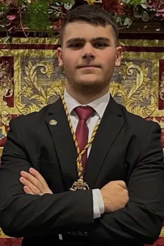Joven con traje oscuro, corbata granate y medalla de Hermandad posa con brazos cruzados frente a un estandarte bordado en dorado y rojo