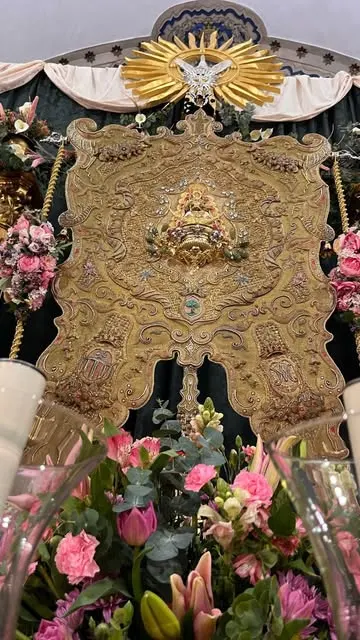 Retablo dorado tallado con el Simpecado en el centro, decorado con flores rosas y verdes y rematado por una paloma blanca sobre un sol radiante
