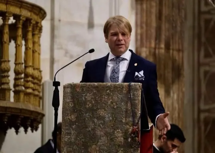 Hombre vestido con traje oscuro y corbata clara hablando en un atril decorado dentro de un edificio histórico con columnas talladas y paredes de piedra clara