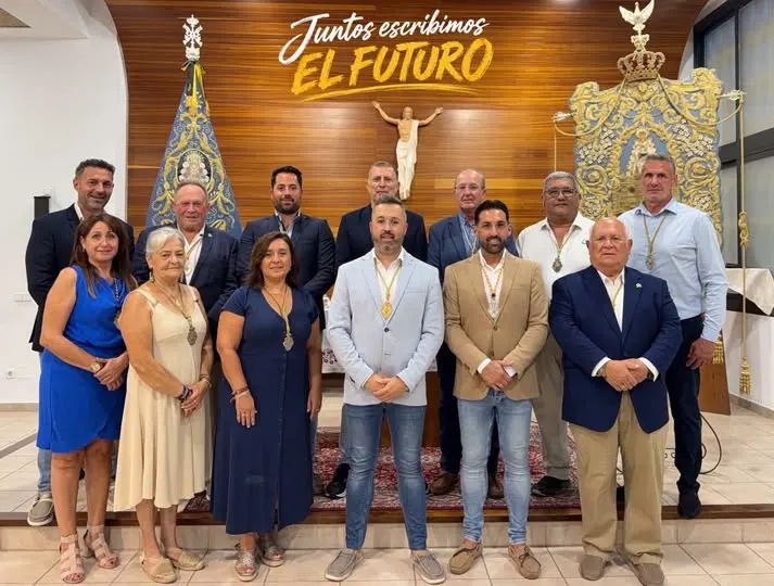 Grupo de trece personas posando en interior con dos estandartes religiosos y un crucifijo sobre fondo de madera con texto Juntos escribimos el futuro