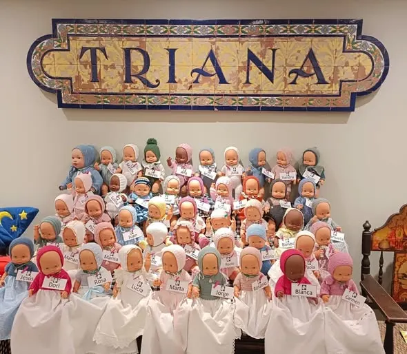 Muñecos vestidos con ropajes tradicionales y etiquetas con nombres, dispuestos en filas bajo un azulejo con la palabra TRIANA en letras grandes y azules