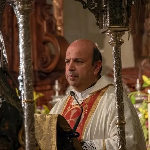 Sacerdote con vestimenta litúrgica blanca y roja oficia ceremonia religiosa en interior de iglesia decorada con ornamentos dorados y madera tallada