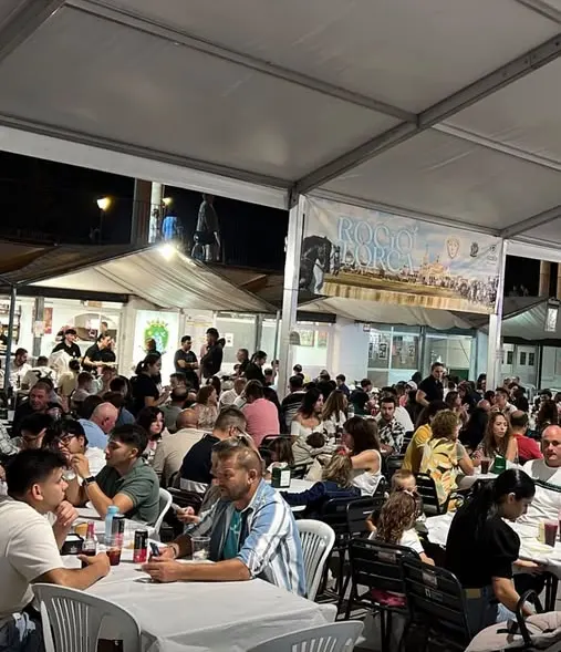 Personas de todas las edades sentadas en mesas bajo una carpa disfrutando de comida y bebida en la caseta de la Hermandad del Rocío de Lorca durante la feria nocturna