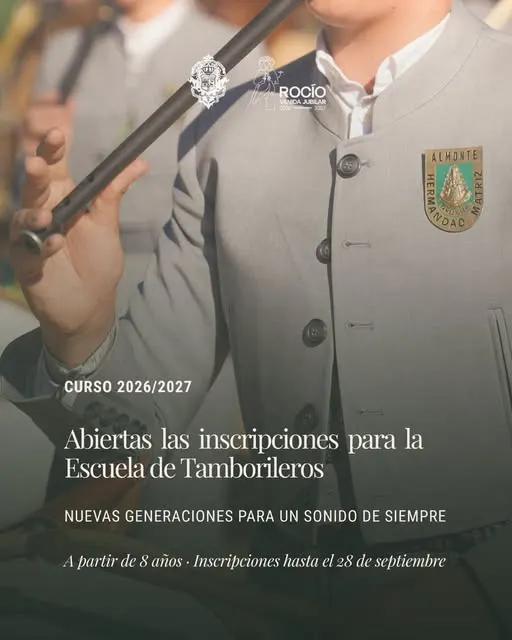 Persona vestida con traje gris claro y escudo de Hermandad Matriz de Almonte tocando un tamboril con baquetas negras