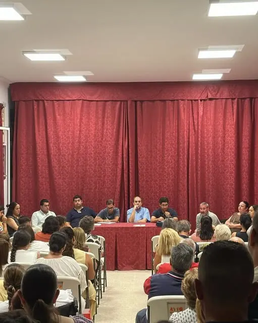 Reunión con asistencia completa donde miembros de la Junta exponen temas electorales y proyectos ante hermanos sentados en sillas frente a mesa presidencial roja
