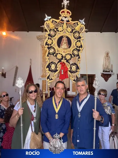Tres personas posan en el interior de una capilla junto a un simpecado decorado con bordados dorados y una imagen religiosa central