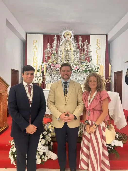 Tres personas vestidas de manera formal posan frente a un altar decorado con flores y una imagen religiosa en un espacio interior sencillo